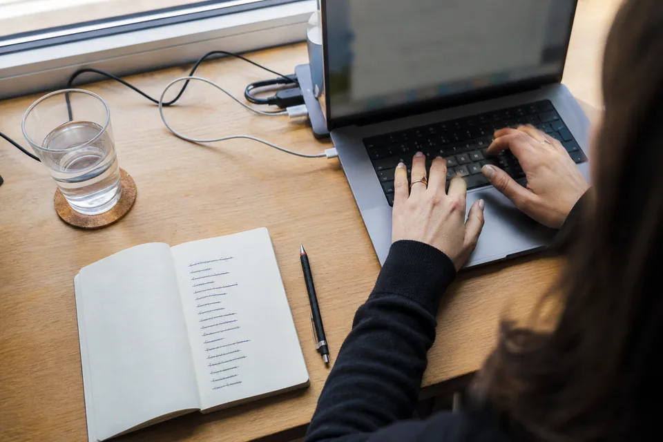 Hands typing on a laptop at a home office desk next to a notebook with a handwritten prospect list