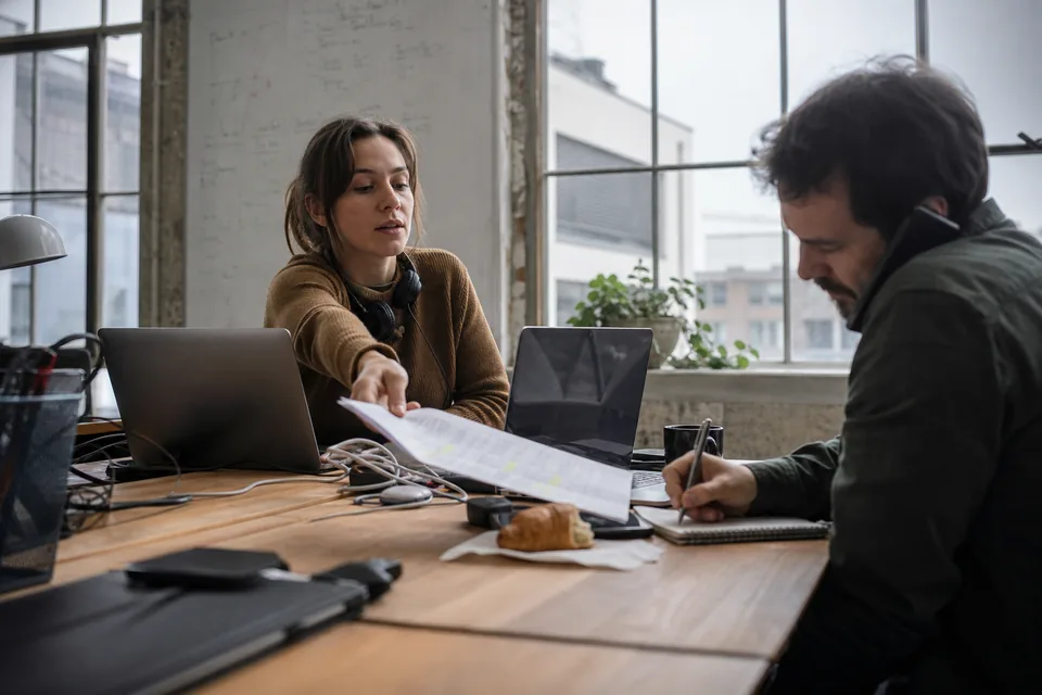 Two sales reps at a shared desk, one sliding a highlighted printed prospect list to a colleague on a call